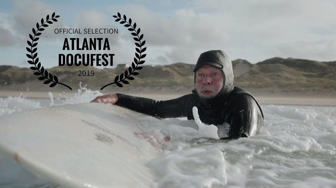 Cover photo of "Solace" the short documentary which shows a man with his surfboard in the water with an official selection laurel from the Atlanta Docufest film festival